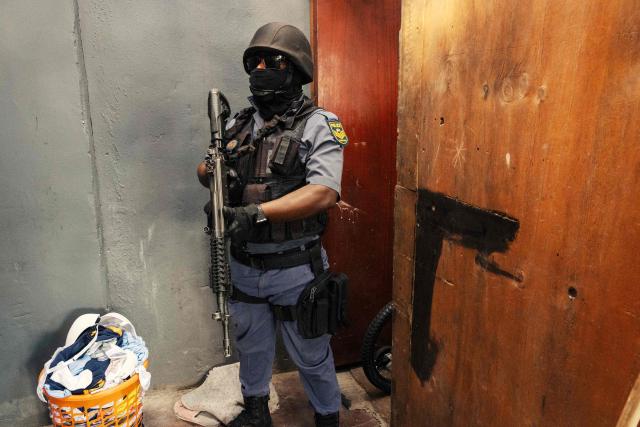 A South African policeman stands guard at the door of house while it is searched during operation Shanela and crime prevention in Tafelsig, Cape Town on February 13, 2026. Operation Shanela, "to sweep" in Setswana, is a high-density, multi-disciplinary, and countrywide crime-fighting initiative led by the South African Police Service (SAPS) to combat serious crime, specifically targeting hotspots, illegal mining, and unlicensed firearms. (Photo by GIANLUIGI GUERCIA / AFP)