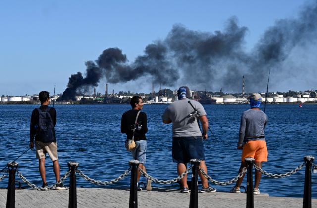Men fish as black smoke billows from a fire at the Nico Lopes oil refinery in Havana on February 13, 2026. A fire broke out at a refinery in the capital of Cuba, already struggling under what amounts to a US blockade of oil deliveries. AFP observed a massive column of smoke rising from the Nico Lopez refinery in Havana Bay, though it was not known if the blaze was near the plant’s oil storage tanks. (Photo by YAMIL LAGE / AFP)