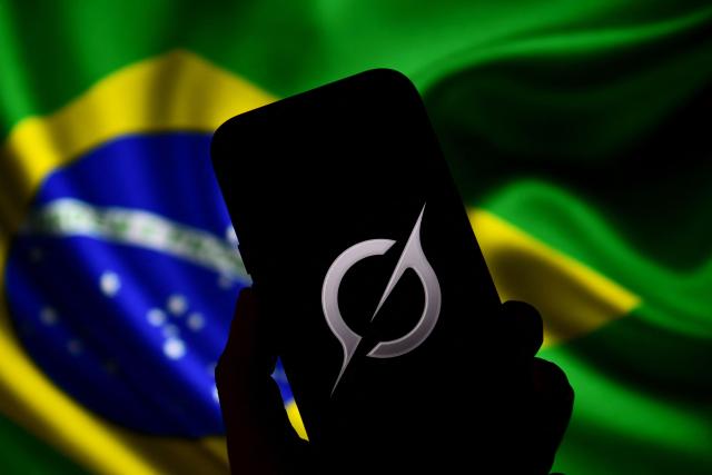 In this photo illustration, the Grok logo is seen on a mobile phone in front of a Brazilian flag displayed on a computer screen in Bogota on February 13, 2026. Brazilian authorities reported on February 12 that they had ordered billionaire Elon Musk's social network X to take immediate action to prevent the creation of sexual images generated by Grok, its artificial intelligence (AI) assistant. (Photo by Pablo VERA / AFP)