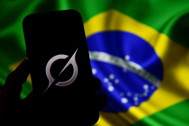 In this photo illustration, the Grok logo is seen on a mobile phone in front of a Brazilian flag displayed on a computer screen in Bogota on February 13, 2026. Brazilian authorities reported on February 12 that they had ordered billionaire Elon Musk's social network X to take immediate action to prevent the creation of sexual images generated by Grok, its artificial intelligence (AI) assistant. (Photo by Pablo VERA / AFP)
