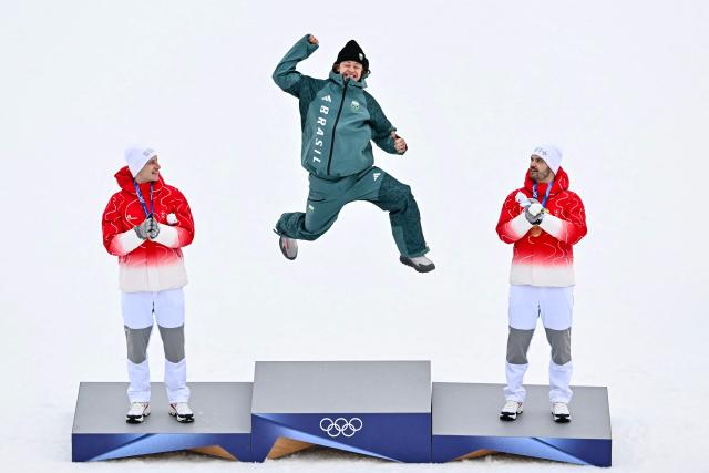 TOPSHOT - Brazil's gold medalist Lucas Pinheiro Braathen (C) leaps onto the podium flanked by Switzerland's silver medalist Marco Odermatt (L), and Switzerland's bronze medalist Loic Meillard during the podium of the men's giant slalom alpine skiing event during the Milano Cortina 2026 Winter Olympic Games at the Stelvio Ski Centre in Bormio (Valtellina) on February 14, 2026. (Photo by Fabrice COFFRINI / AFP)