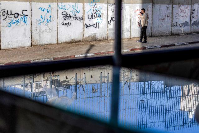 A man speaks on a phone while walking next to Israel's controversial separation barrier, reflected on a surface at a butcher's shop, in the Dahiat al-Barit suburb of east Jerusalem on February 15, 2026. (Photo by Ilia YEFIMOVICH / AFP)