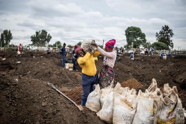 A woman grimaces as she is helped to load a sack of soil on her shoulder in a patch of land where artisanal miners look for gold outside Springs, Ekurhuleni, on February 15, 2026. This land, riddled with holes dug with pickaxes, was just a week ago a cattle pen. Barbed wire still bears witness to this. This patch of land in a Springs township is now attracting gold prospectors from the surrounding area. (Photo by MARCO LONGARI / AFP)