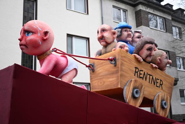 A carnival float designed by German carnival float builder Jacques Tilly featuring a baby pulling a cart with elderly people in it is pictured during the parade to celebrate Rose Monday (Rosenmontag), on February 16, 2026 in Duesseldorf, western Germany. (Photo by INA FASSBENDER / AFP)