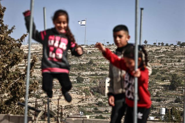Palestinian children play on a trampoline in Umm Safa village, north of Ramallah in the occupied West Bank, opposite an Israeli flag that was raised on a hilltop overlooking the village on February 16, 2026. Israel's government has approved a process to register land in the West Bank as "state property", drawing condemnation from Arab nations and critics who said it would accelerate annexation of the Palestinian territory. (Photo by Zain JAAFAR / AFP)