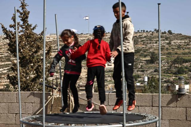 Palestinian children play on a trampoline in Umm Safa village, north of Ramallah in the occupied West Bank, opposite an Israeli flag that was raised on a hilltop overlooking the village on February 16, 2026. Israel's government has approved a process to register land in the West Bank as "state property", drawing condemnation from Arab nations and critics who said it would accelerate annexation of the Palestinian territory. (Photo by Zain JAAFAR / AFP)