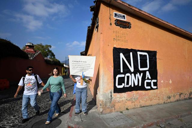 Tourists walk past a protester holding a banner featuring a verse from the poem Noche serena by Spanish poet Fray Luis de Leon outside a hotel as University of San Carlos officials meet to appoint Constitutional Court magistrates, amid demonstrations against the possible selection of Attorney General Consuelo Porras, in Antigua, Guatemala, on February 16, 2026. Guatemala has begun the process to renew members of key judicial institutions, including its top court and the Prosecutor's Office, which for years have been suspected of protecting a network of corruption and criminal activity. (Photo by JOHAN ORDONEZ / AFP)