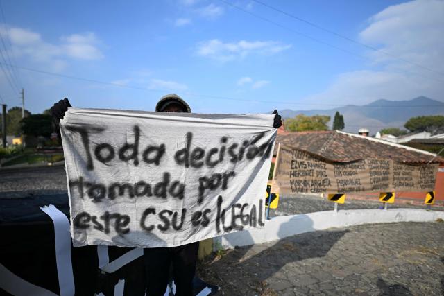 A university student holds a banner reading "Any decision made by this CSU  is illegal"  as University of San Carlos officials meet to appoint Constitutional Court magistrates, amid protests against the possible selection of Attorney General Consuelo Porras, in Antigua, Guatemala, on February 16, 2026. Guatemala has begun the process to renew members of key judicial institutions, including its top court and the Prosecutor's Office, which for years have been suspected of protecting a network of corruption and criminal activity. (Photo by JOHAN ORDONEZ / AFP)