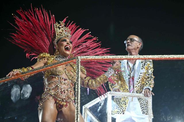 Brazilian drum master Mestre Cica (R) gestures next to a reveller during the parade of the Unidos do Viradouro samba school in his honor on the second night of the Rio Carnival at the Marques de Sapucai Sambadrome in Rio de Janeiro, Brazil, early on February 17, 2026. (Photo by Mauro PIMENTEL / AFP)