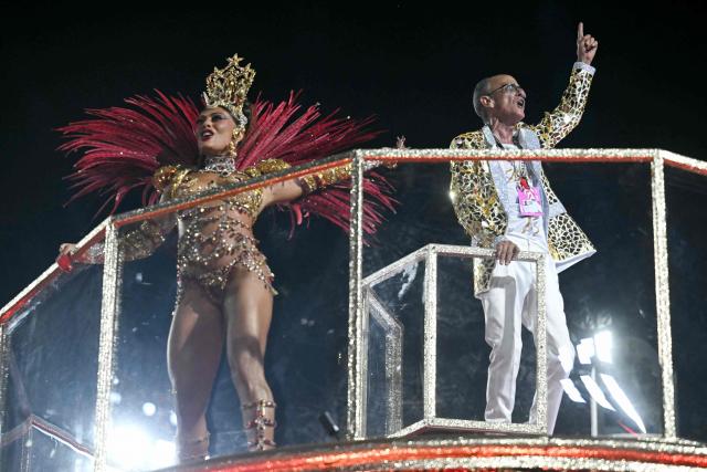 Brazilian drum master Mestre Cica (R) gestures next to a reveller during the parade of the Unidos do Viradouro samba school in his honor on the second night of the Rio Carnival at the Marques de Sapucai Sambadrome in Rio de Janeiro, Brazil, early on February 17, 2026. (Photo by Mauro PIMENTEL / AFP)