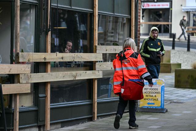 Pedestrians walk past a shop which has been boarded up to protect its frontage, as man sits in the window looking out, ahead of the annual Royal Shrovetide Football match in Ashbourne, northern England on February 17, 2026. The mass-participation ball game involves two teams, whose players are defined by which side of a small brook that bisects the town they were born, aiming to score a goal, which are some three miles (4.8 km) apart. The game, which has very few rules and believed to have been played annually since 1667, is played over two eight hour periods on Shrove Tuesday and Ash Wednesday. (Photo by Paul ELLIS / AFP)