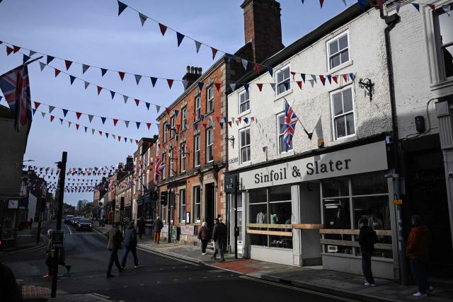 Pedestrians walk past shops which have been boarded up to protect the frontage, as flags decorate the streets above, ahead of the annual Royal Shrovetide Football match in Ashbourne, northern England on February 17, 2026. The mass-participation ball game involves two teams, whose players are defined by which side of a small brook that bisects the town they were born, aiming to score a goal, which are some three miles (4.8 km) apart. The game, which has very few rules and believed to have been played annually since 1667, is played over two eight hour periods on Shrove Tuesday and Ash Wednesday. (Photo by Paul ELLIS / AFP)