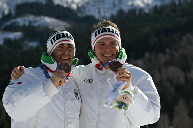 Bronze medallists Italy's Federico Pellegrino (L) and Italy's Elia Barp celebrate on the podium for the men's team cross country free sprint final event of the Milano Cortina 2026 Winter Olympic Games at Tesero Cross-Country Skiing Stadium in Lago di Tesero (Val di Fiemme), on February 18, 2026. (Photo by Javier SORIANO / AFP)