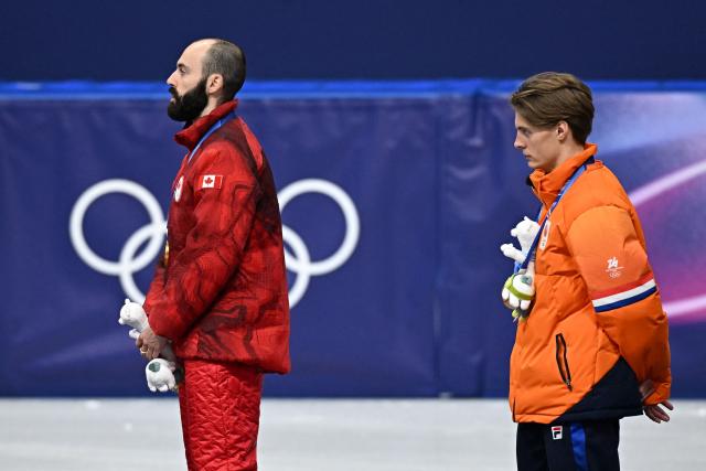 Gold medallist Canada's Steven Dubois listens the Canada's national anthem next to Netherlands' Jens van 't Wout at the podium of the short track speed skating men's 500m final during the Milano Cortina 2026 Winter Olympic Games at Milano Ice Skating Arena in Milan on February 18, 2026. (Photo by Gabriel BOUYS / AFP)