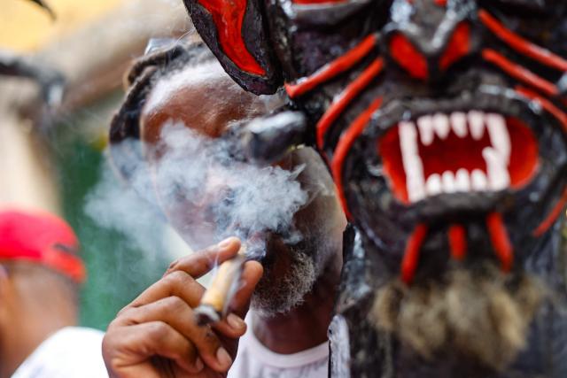 A reveller smokes during the Raimundo Bouzanfraim procession in the Pelourinho district of Salvador, Bahia state, Brazil, on February 18, 2026, marking the beginning of Ash Wednesday and celebrating Afro-Brazilian spirituality and ancestry. This year, the parade celebrates ancestry and Afro-Brazilian spirituality, with the theme of Exu as a symbol of movement, renewal and the connection between past, present and future. (Photo by Rafael Martins / AFP)