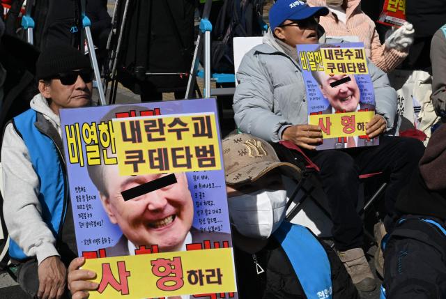A protester holds a placard showing a photo of South Korea's impeached former president Yoon Suk Yeol with words reading "A death sentence" during a rally against Yoon near the Seoul Central District Court in Seoul on February 19, 2026. A South Korean court will rule on February 19, on insurrection charges against ex-president Yoon Suk Yeol, weighing whether he should be sentenced to death for his failed bid to impose military rule. (Photo by Jung Yeon-je / AFP)