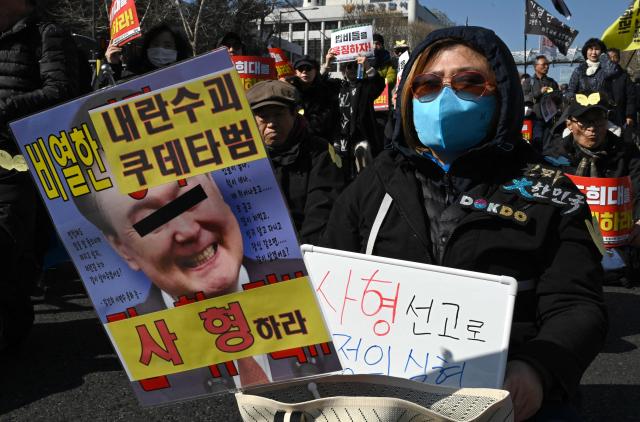 A protester holds a placard showing a photo of South Korea's impeached former president Yoon Suk Yeol with words reading "A death sentence" during a rally against Yoon near the Seoul Central District Court in Seoul on February 19, 2026. A South Korean court will rule on February 19, on insurrection charges against ex-president Yoon Suk Yeol, weighing whether he should be sentenced to death for his failed bid to impose military rule. (Photo by Jung Yeon-je / AFP)