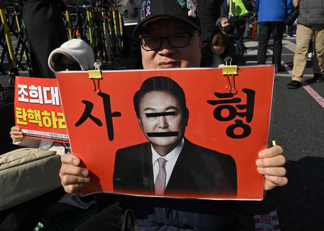 A protester holds a placard showing a photo of South Korea's impeached former president Yoon Suk Yeol reading "A death sentence" during a rally against Yoon near the Seoul Central District Court in Seoul on February 19, 2026. A South Korean court will rule on February 19, on insurrection charges against ex-president Yoon Suk Yeol, weighing whether he should be sentenced to death for his failed bid to impose military rule. (Photo by Jung Yeon-je / AFP)
