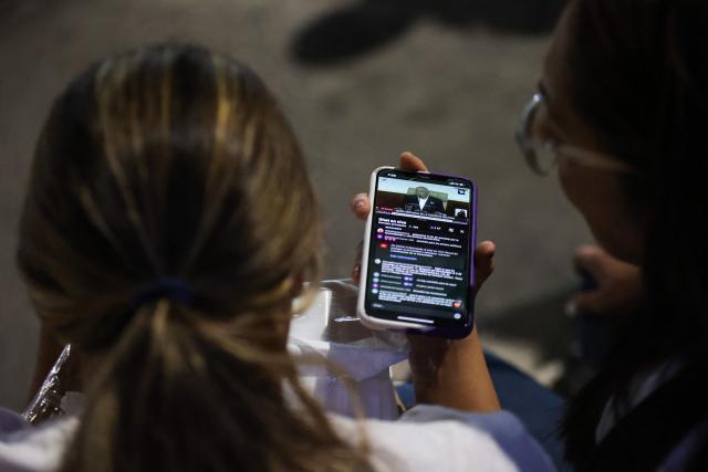 Relatives watch on a cellphone the livestream of Venezuela’s National Assembly President Jorge Rodriguez speaking during a debate on an amnesty bill proposed by Venezuela’s interim president Delcy Rodriguez, outside the El Helicoide building, headquarters of the Bolivarian National Intelligence Service (SEBIN), in Caracas on February 19, 2026. Venezuela's Parliament resumed on February 19 the debate on an amnesty bill that could free hundreds of political prisoners. (Photo by Pedro MATTEY / AFP)