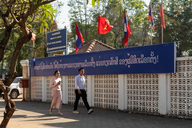 Pedestrians walk past Laos national flags and Lao People's Revolutionary Party flags above a banner that translates as "Voting with respect for the electoral code is voter dignity" ahead of the election in Vientiane on February 19, 2026. The February 22 election will see 243 candidates contest 175 seats after being pre-selected by the ruling communist party. The Southeast Asian nation has no opposition parties, no fully independent news outlets, and the ruling Lao People's Revolutionary Party has held power for more than 50 years. (Photo by AFP)