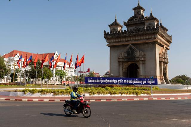 A motorist drives past Laos national flags and Lao People's Revolutionary Party flags and a banner that translates as "Congratulations to the election of representatives to the 10th National Assembly of 5th Members of the Provincial Peoples Councils" displayed ahead of the election next to Patuxai, also known as the Victory Monument, in Vientiane on February 19, 2026. The February 22 election will see 243 candidates contest 175 seats after being pre-selected by the ruling communist party. The Southeast Asian nation has no opposition parties, no fully independent news outlets, and the ruling Lao People's Revolutionary Party has held power for more than 50 years. (Photo by AFP)