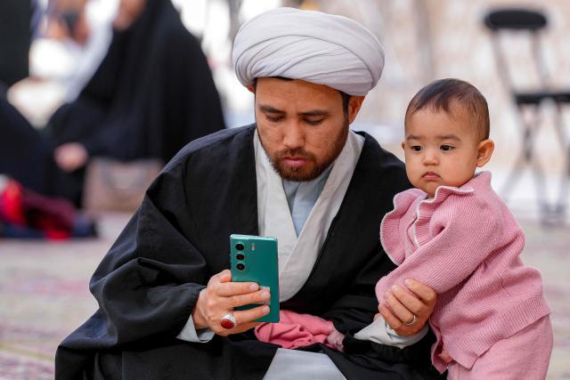 A Shiite Muslim cleric holds a phone as he sits with a toddler in Iraq's central holy city of Najaf on February 19, 2026 during the Muslim fasting month of Ramadan. (Photo by Qassem al-KAABI / AFP)