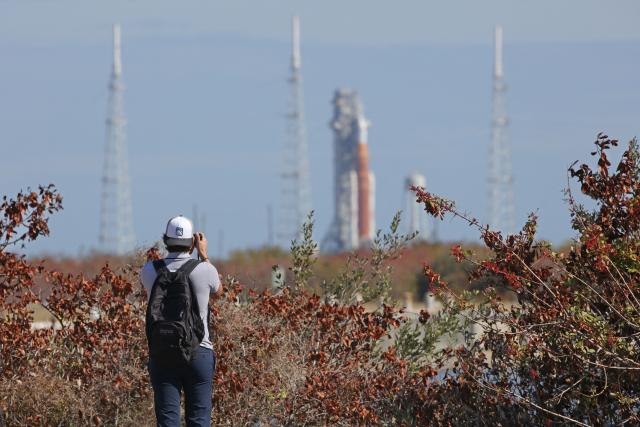 A journalist snaps a photo of NASA's Artemis II Space Launch System (SLS) rocket and Orion spacecraft seen in the distance at Launch Pad 39B at Kennedy Space Center in Cape Canaveral, Florida, on February 20, 2026. NASA performed their second wet dress rehearsal prior to sending four astronauts to the moon for the first time in more than 50 years. NASA officials said they are targeting March 6 for the crewed flight to the moon. (Photo by Gregg Newton / AFP)