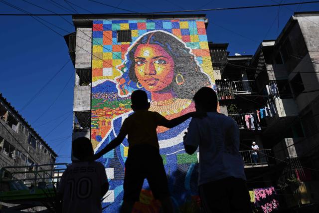Children look at a mural made with plastic bottle caps by Venezuelan artist Oscar Olivares on an apartment building in the Zacamil neighborhood in Mejicanos, El Salvador, on February 20, 2026. The artwork, over 13 meters tall and made with more than 100,000 recycled plastic bottle caps, is regarded as the world's tallest bottle-cap mural. The piece reinterprets the Mona Lisa with a Latin American identity and pays tribute to Salvadoran women. (Photo by Marvin RECINOS / AFP)