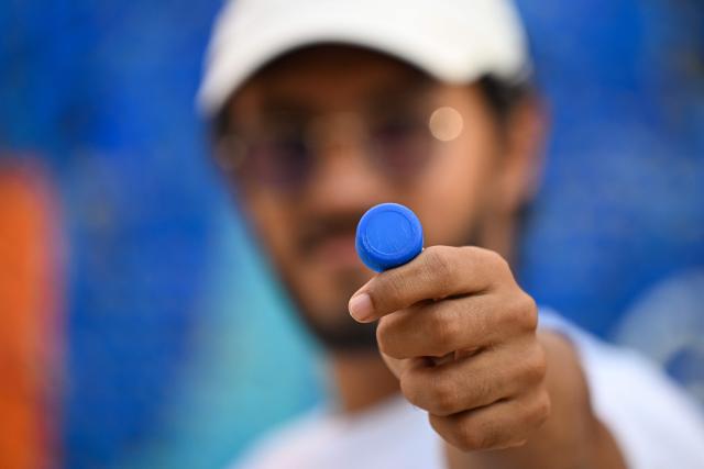 Venezuelan artist Oscar Olivares shows a plastic bottle cap in front of his mural in the Zacamil neighborhood in Mejicanos, El Salvador, on February 20, 2026. The artwork, over 13 meters tall and made with more than 100,000 recycled plastic bottle caps, is regarded as the world's tallest bottle-cap mural. The piece reinterprets the Mona Lisa with a Latin American identity and pays tribute to Salvadoran women. (Photo by Marvin RECINOS / AFP)