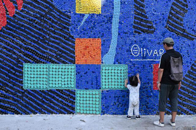 A little girl touches a mural made with plastic bottle caps by Venezuelan artist Oscar Olivares on an apartment building in the Zacamil neighborhood in Mejicanos, El Salvador, on February 20, 2026. The artwork, over 13 meters tall and made with more than 100,000 recycled plastic bottle caps, is regarded as the world's tallest bottle-cap mural. The piece reinterprets the Mona Lisa with a Latin American identity and pays tribute to Salvadoran women. (Photo by Marvin RECINOS / AFP)
