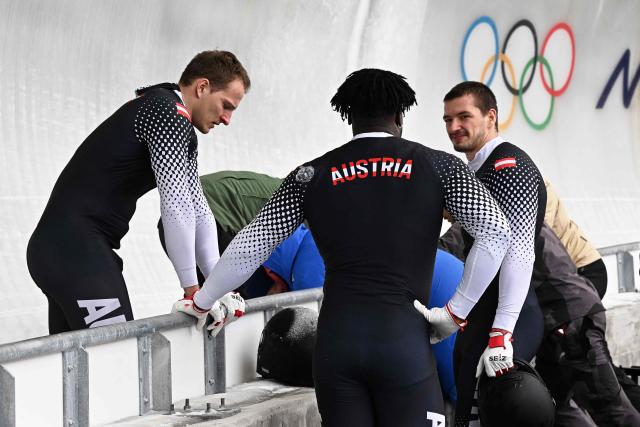 Austria's Daiyehan Nichols-Bardi (C), Austria's Daniel Bertschler (L) and Austria's Sebastian Mitterer (R) react as medics bring off Austria's Jakob Mandlbauer after a crash in the bobsleigh men's 4-man heat 2 at Cortina Sliding Centre during the Milano Cortina 2026 Winter Olympic Games in Cortina d'Ampezzo on February 21, 2026. (Photo by Tiziana FABI / AFP)