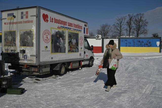 A woman walks past French volunteer baker Loic Nervi mobile bakery that distribute fresh baked bread among residents of a temporary dwelling for people who lost their homes and internally displaced people in Borodyanka, Kyiv region on February 19, 2026, amid the Russian invasion of Ukraine. While Ukraine regularly faces power and heating cuts due to Russian strikes, Loic Nervi works alone from early morning to bake around 700 loaves of bread a day, attracting dozens of locals to his white truck. Over multiple missions across Ukraine, he says he has distributed tens of thousands of loaves, most often to elderly residents left without family support. (Photo by Genya SAVILOV / AFP)