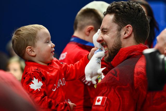 2 1/2 year old Luke (L), son of Canada's Brett Gallant (R) strokes his dads fiace with the Olympic mascot Tina after team Canada defeated team GB in curling at the Milano Cortina 2026 Winter Olympic Games at the Cortina Curling Olympic Stadium in Cortina d’Ampezzo on February 21, 2026. (Photo by Marco BERTORELLO / AFP)