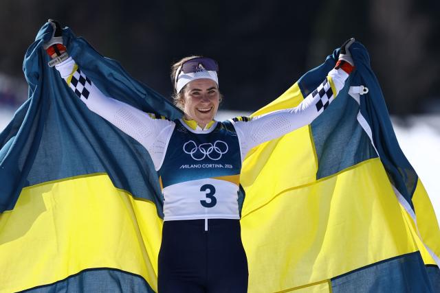 TOPSHOT - Sweden's Ebba Andersson celebrates with a flag of Sweden after crossing the finish line to win the women's cross country 50km mass start final event of the Milano Cortina 2026 Winter Olympic Games at Tesero Cross-Country Skiing Stadium in Lago di Tesero (Val di Fiemme), on February 22, 2026. (Photo by Anne-Christine POUJOULAT / AFP)