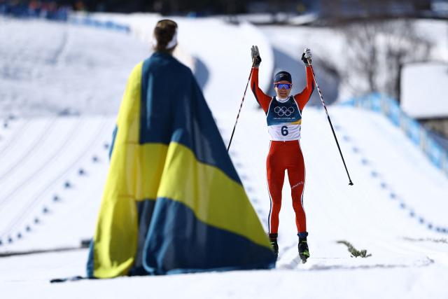 Sweden's Ebba Andersson celebrates with a flag of Sweden (L) next to Norway's Heidi Weng at the end of the women's cross country 50km mass start final event of the Milano Cortina 2026 Winter Olympic Games at Tesero Cross-Country Skiing Stadium in Lago di Tesero (Val di Fiemme), on February 22, 2026. (Photo by Anne-Christine POUJOULAT / AFP)