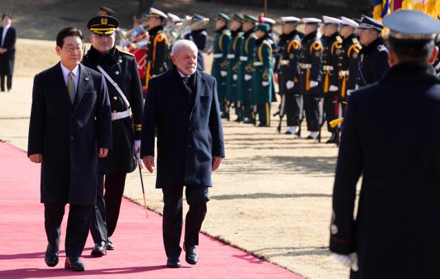 Brazil's President Luiz Inacio Lula da Silva (C) and  South Korea's President Lee Jae Myung (L) inspect an honour guard during a welcome ceremony at the presidential Blue House in Seoul on February 23, 2026. Brazilians could more easily get their hands on coveted "K-beauty" products under a deal struck on February 23 during President Luiz Inacio Lula da Silva's state visit to South Korea. Lula and South Korean leader Lee Jae Myung signed a slew of agreements with the Brazilian president in Seoul, covering everything from agriculture to business cooperation. (Photo by JEON HEON-KYUN / POOL / AFP)
