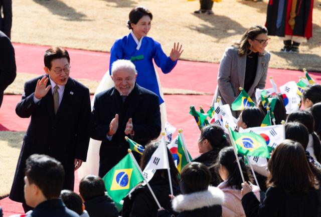 Brazil's President Luiz Inacio Lula da Silva (2nd L) and his wife Rosangela da Silva (R), South Korea's President Lee Jae Myung (L) and his wife Kim Hea Kyung (3rd L) greet children waving South Korean and Brazilian national flags during a welcome ceremony at the presidential Blue House in Seoul on February 23, 2026. Brazilians could more easily get their hands on coveted "K-beauty" products under a deal struck on February 23 during President Luiz Inacio Lula da Silva's state visit to South Korea. Lula and South Korean leader Lee Jae Myung signed a slew of agreements with the Brazilian president in Seoul, covering everything from agriculture to business cooperation. (Photo by JEON HEON-KYUN / POOL / AFP)
