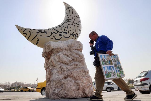 An ambulant photographer speaks on a phone as he walks past a sculpture depicting a crescent moon covered with inscriptions of the Islamic names of God, set up for the Holy month of Ramadan, in Tehran on February 23, 2026. (Photo by ATTA KENARE / AFP)