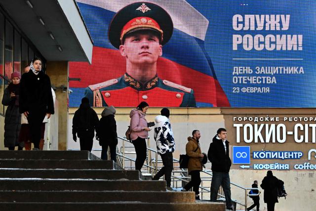 People walk past a giant digital screen displaying an image of a Russian soldier with the slogan "(I) serve Russia!" on Defender of the Fatherland Day in Saint Petersburg on February 23, 2026. (Photo by Olga MALTSEVA / AFP)