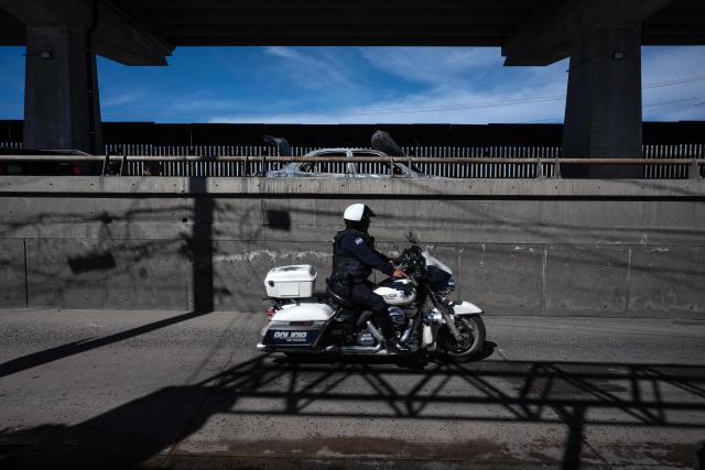 A Mexican police officer drives a motorbike past a burned car, allegedly set on fire by organized crime groups, on International Avenue on the Mexican side of the US-Mexico border in Tijuana, Baja California State, Mexico on February 23, 2026. Mexico has deployed 10,000 troops to quell clashes sparked by the killing of the country's most-wanted drug lord, Nemesio "El Mencho" Oseguera, leader of the Jalisco New Generation Cartel (CJNG), that have claimed dozens of lives, officials said on February 23. News of his death triggered spasms of violence, with cartel members blocking roads in 20 states and torching vehicles and businesses. (Photo by Guillermo ARIAS / AFP)