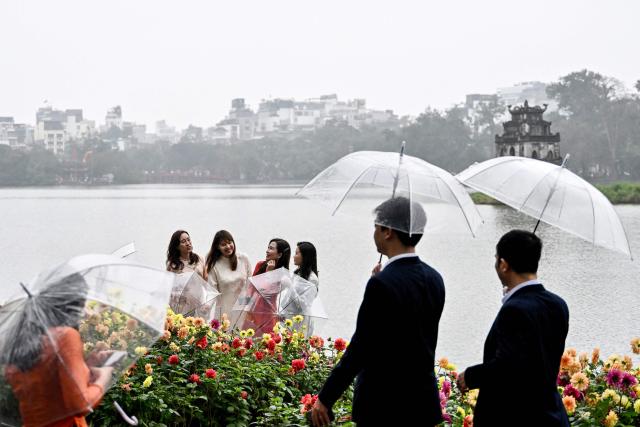People walk holding umbrellas as women pose for photos beside Hoan Kiem Lake in Hanoi on February 24, 2026. (Photo by Nhac NGUYEN / AFP)