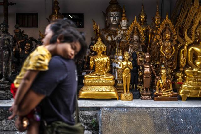 A pedestrian carrying a child walks past a shop selling Buddha statues in Bangkok on February 24, 2026. (Photo by chanakarn laosarakham / AFP)