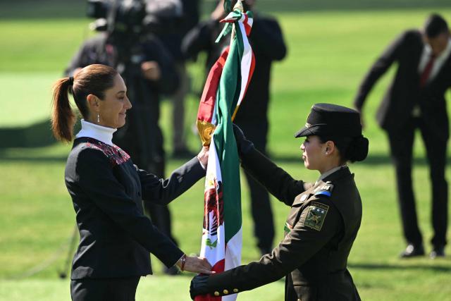 Mexico’s President Claudia Sheinbaum (L) receives a Mexican flag from a member of the Mexican Army during the celebration of Flag Day in Mexico City on February 24, 2026. Sheinbaum offered "full guarantees" for the safety of football fans at World Cup games in the violence-plagued city of Guadalajara on February 24. (Photo by YURI CORTEZ / AFP)