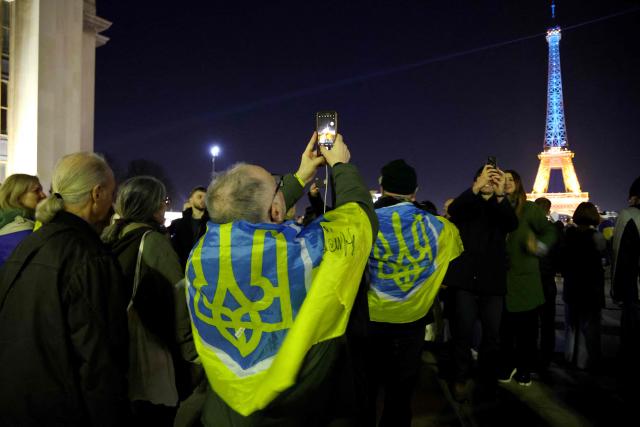 A man draped in a Ukrainian flag takes a photograph of the Eiffel tower as it is illumated in the colours of the Ukrainian flag to mark the fourth anniversary of Russia's war of aggression against Ukraine in Paris on February 24, 2026. Russia launched its full-scale invasion of Ukraine on February 24, 2022, unleashing the deadliest war in Europe since World War II. (Photo by Ludovic MARIN / AFP)