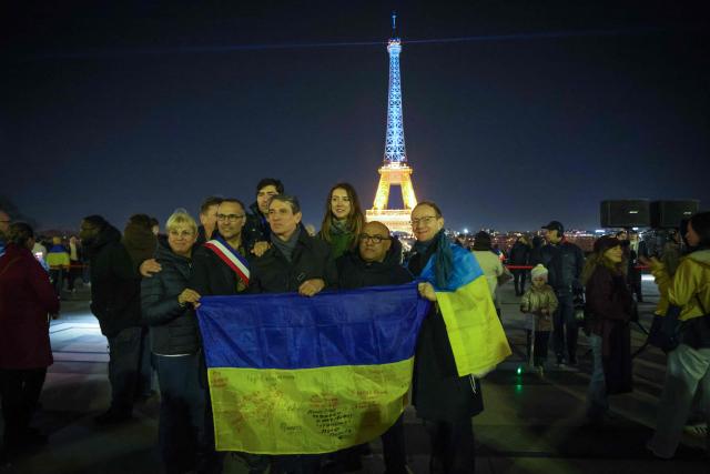 A group pose with a Ukrainian flag in front of the Eiffel tower as it is illumated in the colours of the Ukrainian flag to mark the fourth anniversary of Russia's war of aggression against Ukraine, in Paris on February 24, 2026. Russia launched its full-scale invasion of Ukraine on February 24, 2022, unleashing the deadliest war in Europe since World War II. (Photo by Ludovic MARIN / AFP)