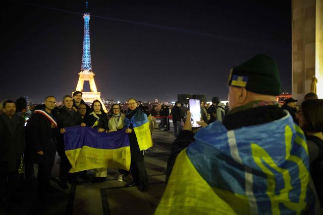 A group pose with a Ukrainian flag in front of the Eiffel tower as it is illumated in the colours of the Ukrainian flag to mark the fourth anniversary of Russia's war of aggression against Ukraine, in Paris on February 24, 2026. Russia launched its full-scale invasion of Ukraine on February 24, 2022, unleashing the deadliest war in Europe since World War II. (Photo by Ludovic MARIN / AFP)