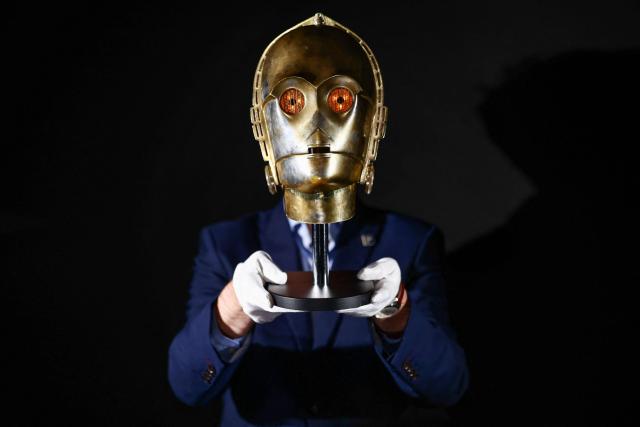 This photo taken on January 30, 2026 shows a gallery assistant posing with a 'Light-Up C-3PO Head' from the 1980 movie "Star Wars: The Empire Strikes Back", at a preview by memorabilia auction house Propstore in Rickmansworth, near London. A collection of around 1,550 pieces of film and TV history will go under the hammer over three days from March 25, with in-room bidding on the first day only at the Petersen Automotive Museum in Los Angeles. (Photo by HENRY NICHOLLS / AFP)