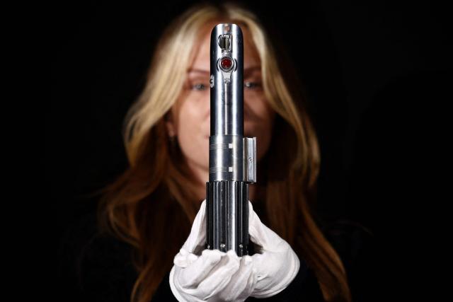 This photo taken on January 30, 2026 shows a gallery assistant posing with 'Rey and Luke Skywalker’s Hero Lightsaber' from the 2015 movie "Star Wars: The Force Awakens", at a preview by memorabilia auction house Propstore in Rickmansworth, near London. A collection of around 1,550 pieces of film and TV history will go under the hammer over three days from March 25, with in-room bidding on the first day only at the Petersen Automotive Museum in Los Angeles. (Photo by HENRY NICHOLLS / AFP)