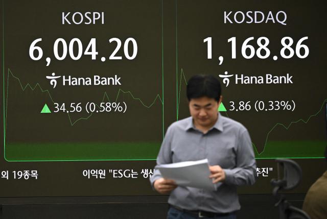 A currency dealer walks past an electronic screen showing South Korea's benchmark stock index (L) and the Korean Securities Dealers Automated Quotations (R) in a foreign exchange dealing room at the Hana Bank headquarters in Seoul on February 25, 2026. South Korea's benchmark Kospi index topped 6,000 points for the first time on February 25, buoyed by the strong performance of the nation's world-leading semiconductor industry. (Photo by Jung Yeon-je / AFP)