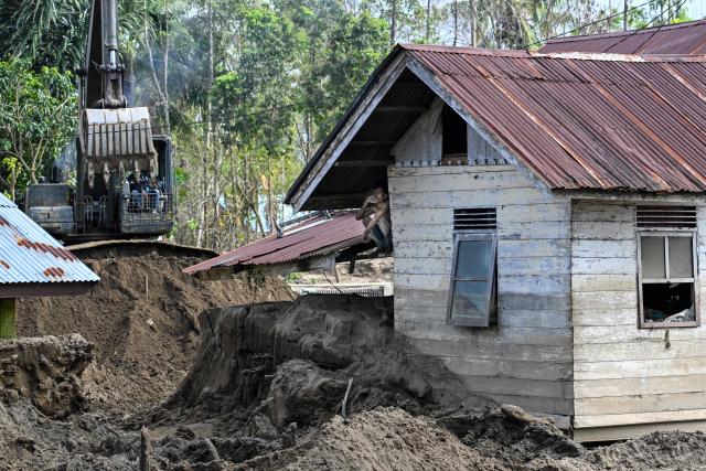 A man (C) sits under the roof of a flood-damaged house while an excavator (L) digs out hardened mud in Meurah Dua, Aceh province's Pidie Jaya district on February 21, 2026, after devastating floods and landslides struck Indonesia's Sumatra late last year. Some 32,000 flood survivors remain displaced following the late 2025 monsoon deluge. In hardest-hit Aceh province, which accounted for most of the more than 1,000 deaths, families have become increasingly frustrated over authorities' sluggish response. Many remain crammed in temporary shelters or a cluster of tents, with little expectation of returning to their mud-caked homes any time soon. (Photo by CHAIDEER MAHYUDDIN / AFP) / To go with AFP story Indonesia-flood by Alfath Asmunda